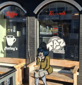 A cartoon woman with blue hair holds a cup of coffee, sitting on a bench outside Morley's Coffee and Taz Records storefront, lost in her own story.